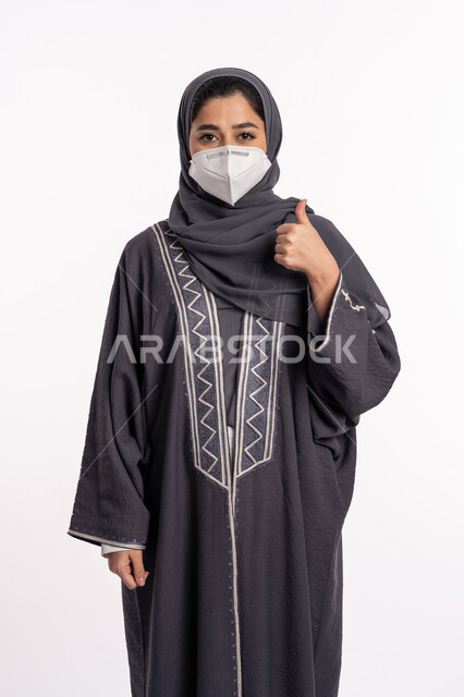 اتباع الإجراءات الاحترازية للوقائية من فايروس كورونا ،الشعور بالقبول والرضا, بورتريه مقرب لإمرأة عربية إماراتية خليجية محجبة ترتدي الكمامة ترفع اصبع الإبهام للأعلى بعلامة الجودة, الحفاظ على السلامة و تجنب انتشار العدوى من كوفيد 19، خلفية بيضاء