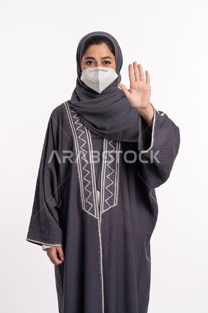 تجنب انتشار العدوى من كوفيد 19، الحفاظ على السلامة، اتباع الإجراءات الاحترازية للوقائية من فايروس كورونا، الرفض ومنع الاقتراب, بورتريه لإمرأة عربية إماراتية خليجية محجبة ترتدي الكمامة ترفع كف يدها لأعلى بتعابير التحذير والتوقف, خلفية بيضاء