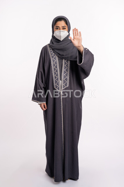 تجنب انتشار العدوى من كوفيد 19، الحفاظ على السلامة، اتباع الإجراءات الاحترازية للوقائية من فايروس كورونا، الرفض ومنع الاقتراب, بورتريه لإمرأة عربية إماراتية خليجية محجبة ترتدي الكمامة ترفع كف يدها لأعلى بتعابير التحذير والتوقف, صورة الجسم بالكامل، خلفية بيضاء