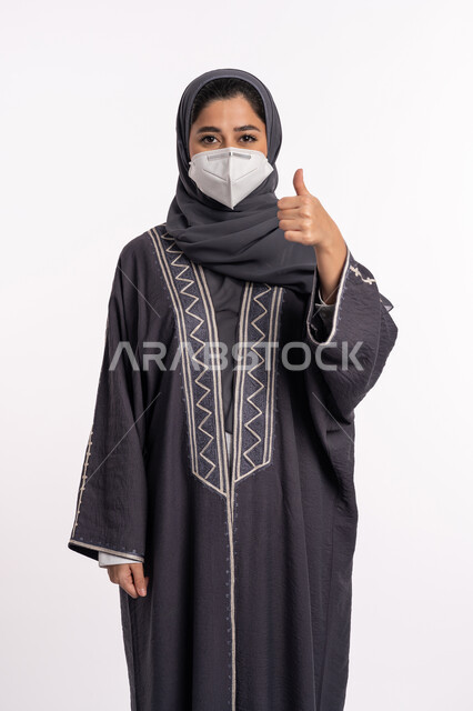 اتباع الإجراءات الاحترازية للوقائية من فايروس كورونا ،الشعور بالقبول والرضا, بورتريه مقرب لإمرأة عربية إماراتية خليجية محجبة ترتدي الكمامة ترفع اصبع الإبهام للأعلى بعلامة الجودة, الحفاظ على السلامة و تجنب انتشار العدوى من كوفيد 19، خلفية بيضاء