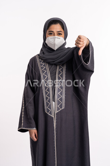 Dislike and approval, avoid the spread of infection from Covid 19, maintain safety, follow precautionary measures to prevent the Corona virus, portrait of a veiled Arab-Emirati Gulf woman wearing a mask, pointing with a thumb down, white background