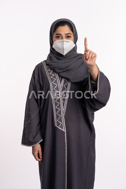 الاشارة الى شيء ما, تجنب انتشار العدوى من كوفيد 19، الحفاظ على السلامة، اتباع الإجراءات الاحترازية للوقائية من فايروس كورونا، بورتريه لإمرأة عربية إماراتية خليجية محجبة ترتدي الكمامة تشير بأصبع السبابة الى الاعلى, خلفية بيضاء