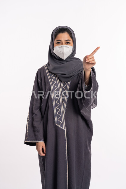 الاشارة الى شيء ما, تجنب انتشار العدوى من كوفيد 19، الحفاظ على السلامة، اتباع الإجراءات الاحترازية للوقائية من فايروس كورونا، بورتريه لإمرأة عربية إماراتية خليجية محجبة ترتدي الكمامة تشير بأصبع السبابة الى الاعلى, خلفية بيضاء