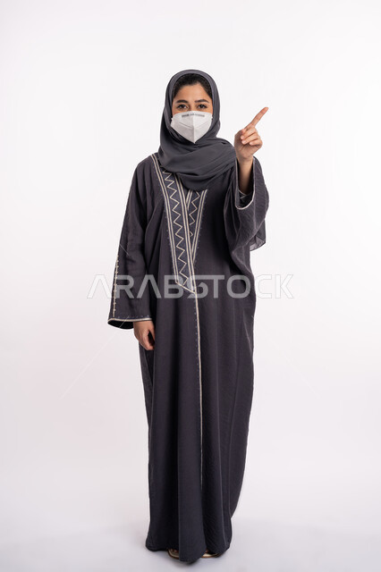 Pointing to something, avoiding the spread of infection from Covid 19, maintaining safety, following precautionary measures to prevent the Corona virus, portrait of a veiled Arab Emirati Gulf woman wearing a mask, pointing with her index finger upward, full body portrait, white background