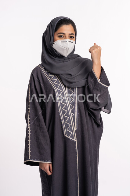 Expressions and gestures of strength and challenge, avoiding the spread of infection from Covid 19, maintaining safety, following precautionary measures to prevent the Corona virus, a portrait of a veiled Arab Emirati Gulf woman wearing a mask, raising her fist upwards, white background.