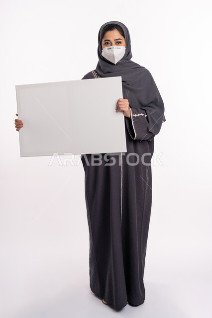 لوحات توعوية لتجنب إنتشار العدوى، الحفاظ على السلامة من كوفيد 19، اتباع الاجراءات الاحترازية للوقاية من فايروس كورونا، بورتريه لإمرأة عربية خليجية إماراتية محجبة ترتدي الكمامة تمسك بيدها موك اب مستطيل أبيض فارغ, صورة الجسم بالكامل، خلفية بيضاء