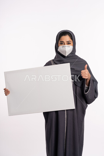 موك اب أبيض فارغ، الشعور بالقبول والرضا، اتباع الاجراءات الاحترازية للوقاية من فايروس كورونا، بورتريه لإمرأة عربية خليجية إماراتية محجبة ترتدي الكمامة تمسك بيدها لوحات توعوية لتجنب إنتشار العدوى ترفع أصبع الأبهام بعلامة الجودة, خلفية بيضاء