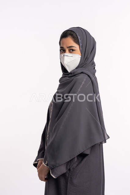 الحفاظ على السلامة من كوفيد 19، اتباع الاجراءات الاحترازية للوقاية من فايروس كورونا، بورتريه مقرب من الجانب لإمرأة عربية خليجية إماراتية محجبة ترتدي الكمامة تقف بإيماءات أيدي متماسكة وتنظر إلى الكاميرا, الوعي الصحي لتجنب إنتشار العدوى، خلفية بيضاء