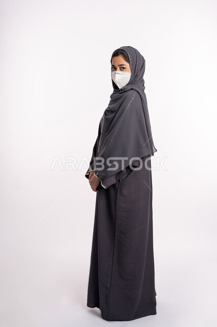 الوعي الصحي لتجنب إنتشار العدوى، الحفاظ على السلامة من كوفيد 19، اتباع الاحترازات الوقائية من فايروس كورونا، بورتريه من الجانب لإمرأة عربية خليجية إماراتية محجبة ترتدي الكمامة تقف بإيماءات أيدي متماسكة وتنظر إلى الكاميرا, صورة الجسم بالكامل، خلفية بيضاء