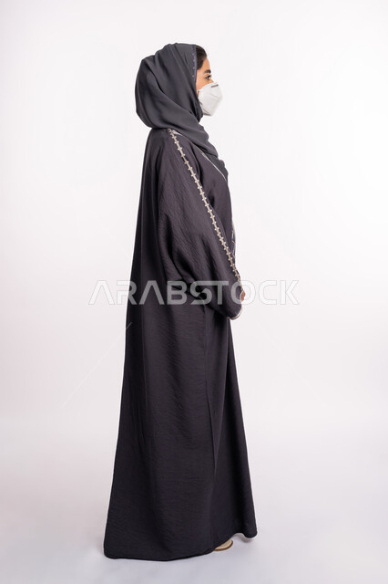 الوعي الصحي لتجنب إنتشار العدوى، الحفاظ على السلامة من كوفيد 19، اتباع الاحترازات الوقائية من فايروس كورونا، بورتريه من الجانب لإمرأة عربية خليجية إماراتية محجبة ترتدي الكمامة تقف بإيماءات أيدي متماسكة وتنظر إلى شيء ما, صورة الجسم بالكامل، خلفية بيضاء