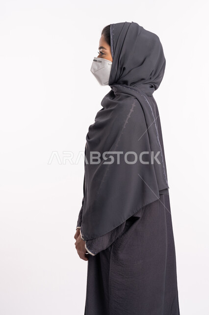اتباع الاجراءات الاحترازية للوقاية من فايروس كورونا، بورتريه مقرب من الجانب لإمرأة عربية خليجية إماراتية محجبة ترتدي الكمامة تقف بإيماءات أيدي متماسكة وتنظر إلى شيء ما, الوعي الصحي لتجنب إنتشار العدوى، الحفاظ على السلامة من كوفيد 19، خلفية بيضاء