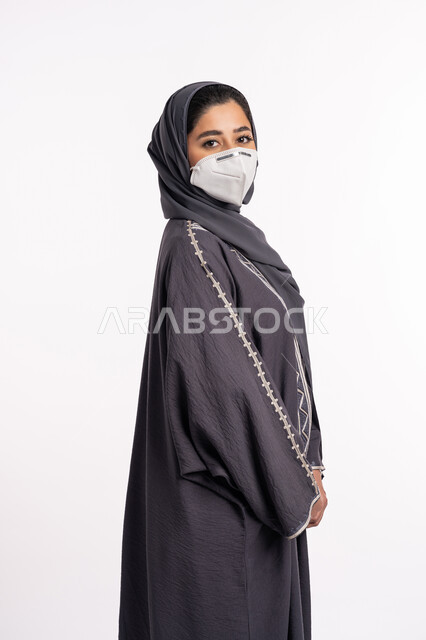 الحفاظ على السلامة من كوفيد 19، اتباع الاجراءات الاحترازية للوقاية من فايروس كورونا، بورتريه مقرب من الجانب لإمرأة عربية خليجية إماراتية محجبة ترتدي الكمامة تقف بإيماءات أيدي متماسكة وتنظر إلى الكاميرا, الوعي الصحي لتجنب إنتشار العدوى، خلفية بيضاء