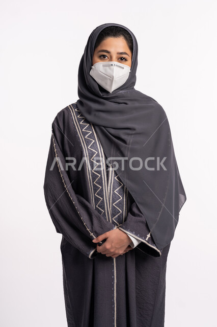 اتباع الاجراءات الاحترازية للوقاية من فايروس كورونا، بورتريه مقرب لإمرأة عربية خليجية إماراتية محجبة ترتدي الكمامة تقف بإيماءات أيدي متماسكة وتنظر إلى الكاميرا, الوعي الصحي لتجنب إنتشار العدوى، الحفاظ على السلامة من كوفيد 19، خلفية بيضاء