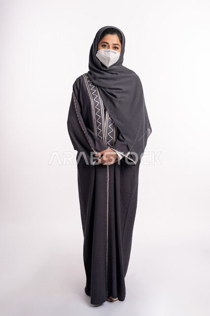 الوعي الصحي لتجنب إنتشار العدوى، الحفاظ على السلامة من كوفيد 19، اتباع الإجراءات الاحترازية للوقائية من فايروس كورونا، بورتريه لإمرأة عربية خليجية إماراتية محجبة ترتدي الكمامة تقف بإيماءات أيدي متماسكة وتنظر إلى الكاميرا , صورة الجسم بالكامل، خلفية بيضاء