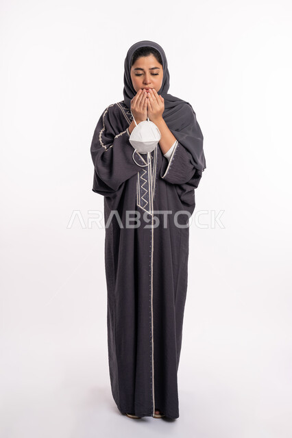 Praying to God for a speedy recovery, following preventive measures against the Corona virus, portrait of an Arab Gulf Emirati woman wearing the abaya and hijab, holding a mask in her hand, praying to God with expressions and gestures of sadness and despair, health awareness to avoid the spread of infection, full body portrait, white background