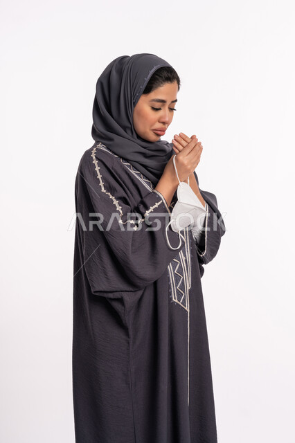 Praying to God for a speedy recovery, following precautionary measures to prevent the Corona virus, a portrait of an Arab Gulf Emirati woman wearing the abaya and hijab, holding a mask in her hand, praying to God with expressions and gestures of sadness and despair, health awareness to avoid the spread of infection, white background.