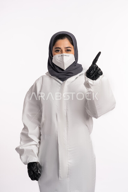 الاشارة الى شيء ما, تجنب انتشار العدوى من كوفيد 19، الحفاظ على السلامة، اتباع الإجراءات الاحترازية للوقائية من فايروس كورونا، بورتريه لإمرأة عربية إماراتية خليجية محجبة ترتدي الرداء الطبي الواقي والقفازات تضع الكمامة تشير بأصبع السبابة الى الاعلى, خلفية بيضاء