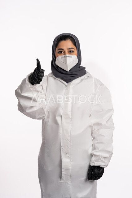 الشعور بالقبول والرضا، اتباع الإجراءات الوقائية من فايروس كورونا, بورتريه لإمرأة عربية إماراتية خليجية محجبة ترتدي الرداء الطبي الواقي والقفازات تضع الكمامة ترفع اصبع الإبهام للأعلى بعلامة الجودة, الحفاظ على السلامة و تجنب انتشار العدوى من كوفيد 19، خلفية بيضاء