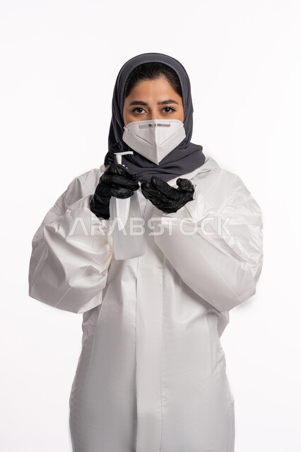 تعقيم اليدين باستخدام المواد الكيميائية والكحول للقضاء على الفايروسات والجراثيم، اتباع الاجراءات الاحترازية للوقاية من فايروس كورونا، بورتريه لإمرأة عربية إماراتية خليجية محجبة ترتدي الرداء الطبي الواقي والقفازات تضع الكمامة تمسك علبة المعقم, خلفية بيضاء