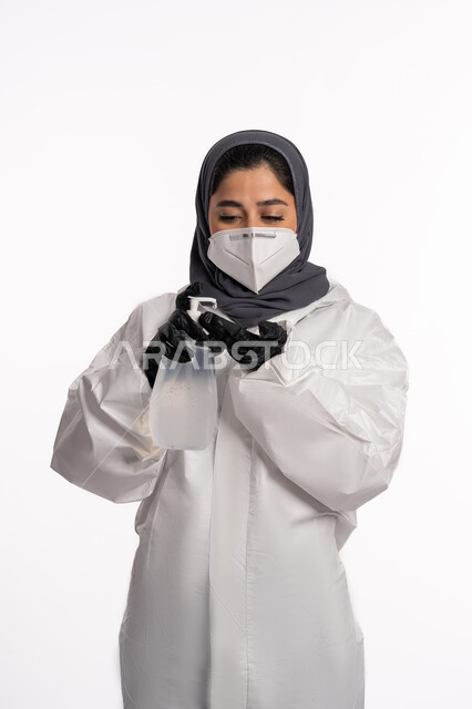 تعقيم اليدين باستخدام المواد الكيميائية والكحول للقضاء على الفايروسات والجراثيم، اتباع الاجراءات الاحترازية للوقاية من فايروس كورونا، بورتريه لإمرأة عربية إماراتية خليجية محجبة ترتدي الرداء الطبي الواقي والقفازات تضع الكمامة تمسك علبة المعقم, خلفية بيضاء