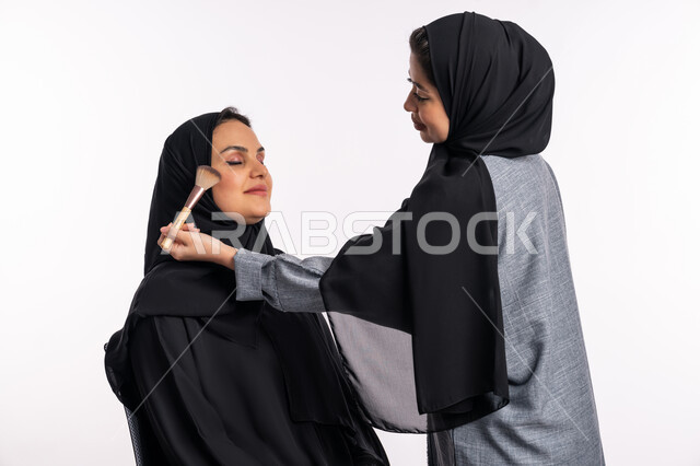 The concept of caring for beauty, skin, and external appearance, a Saudi expert in women’s cosmetics, a close-up portrait of an Arab Gulf Emirati woman wearing an abaya and hijab, applying blusher to brighten the cheeks of her friend, educational courses for tutorial make-up, white background
