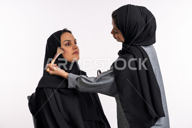 مفهوم الإهتمام بالجمال والبشرة والمظهر الخارجي، خبيرة سعودية لمستحضرات التجميل النسائية، بورتريه مقرب لإمرأة عربية خليجية إماراتية ترتدي العباءة والحجاب تقوم بوضع البلاشر مورد الخدود لصديقتها، دورات تعليمية لمكياج تتوريال، خلفيه بيضاء