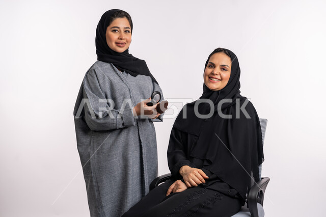 Tutorial makeup courses, the concept of caring for beauty, skin, and external appearance, a Saudi expert in women’s cosmetics, a close-up portrait of an Arab Gulf-Emirati woman wearing the abaya and hijab applying makeup for her friend, white background