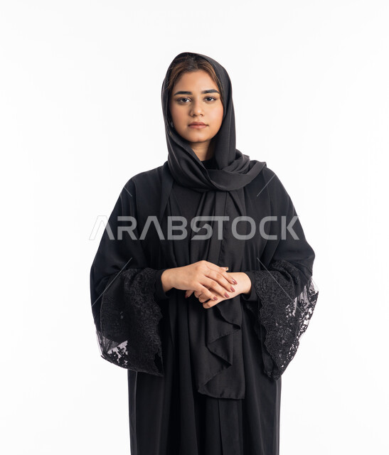 تعابير الثقة بالنفس، شابة سعودية تنظر إلى الكاميرا بايماءات الحدة و الجدية، الاهتمام بالمظهر الخارجي, بورتريه مقرب لامرأة عربية خليجية اماراتية محجبة ترتدي عباءة سوداء تقف بأيدي متشبكة، خلفية بيضاء