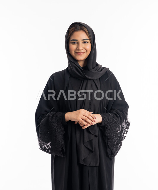 التزيين و التجمل والاهتمام بالمظهر الخارجي، شابة سعودية تنظر إلى الكاميرا بايماءات السرور و الرضى، بورتريه مقرب لإمراة عربية اماراتية خليجية ترتدي العباءة والحجاب تقف باستقامة وثقة النفس، خلفية بيضاء