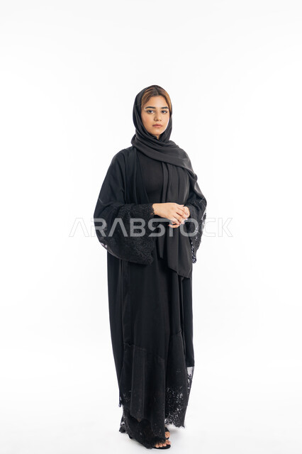 الوقوف باستقامة والاعتزاز بالنفس, الاهتمام بالمظهر الخارجي, شابة سعودية تنظر إلى الكاميرا بايماءات الحزم والجدية، بورتريه مقرب لامرأة عربية خليجية اماراتية محجبة ترتدي عباءة سوداء تقف بأيدي متشبكة، الجسم بالطول الكامل، خلفية بيضاء
