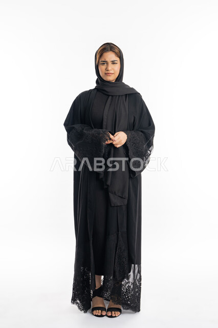 Gestures of anger and nervousness, concern for external appearance, a young Saudi woman looking at the camera with firm gestures, portrait of a veiled Arab Gulf Emirati woman wearing a black abaya, standing with clasped hands, full-length body, white background