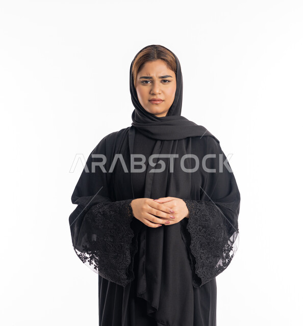تعابير الجدية والانزعاج, الاهتمام بالمظهر الخارجي, شابة سعودية تنظر إلى الكاميرا بايماءات الحزم ، بورتريه مقرب لامرأة عربية خليجية اماراتية محجبة ترتدي عباءة سوداء تقف بأيدي متشبكة، خلفية بيضاء