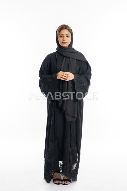 مفهوم الأنوثة والنعومة، الوقوف بجدية وعدم رضا, الاهتمام بالمظهر الخارجي, شابة سعودية تنظر إلى الاسفل بايماءات الحزم والجدية، بورتريه لامرأة عربية خليجية اماراتية محجبة ترتدي عباءة سوداء تقف بأيدي متشبكة، الجسم بالطول الكامل، خلفية بيضاء