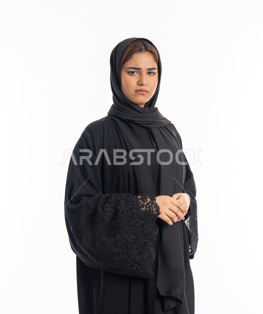 مفهوم الاناقة والانوثة، تعابير الثقة بالنفس، الاهتمام بالمظهر الخارجي, شابة سعودية تنظر إلى الكاميرا بايماءات الحزم والجدية،  بورتريه مقرب لامرأة عربية خليجية اماراتية محجبة ترتدي عباءة سوداء تقف بأيدي متشبكة، خلفية بيضاء