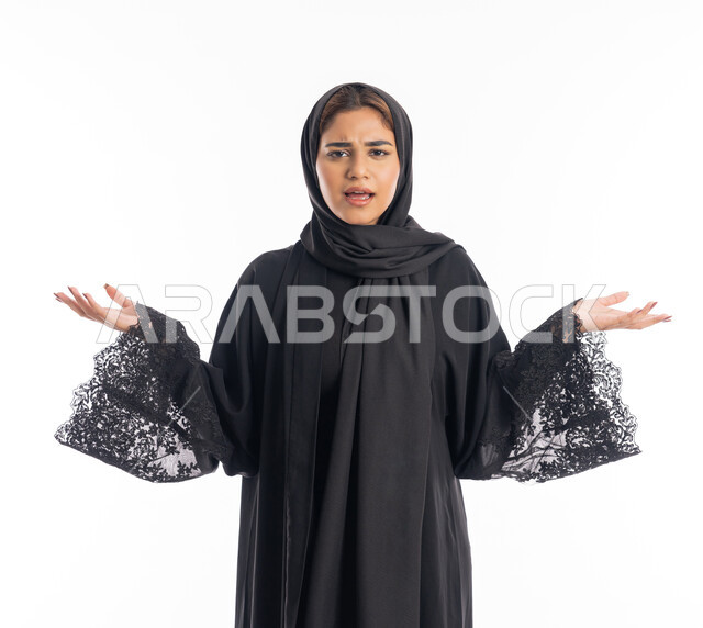 المفاجأة و الاندهاش من شيء ما، شابة سعودية تنظر الى الكاميرا بإستغراب، مفهوم النعومة والانوثة والاناقة، بورتريه مقرب لإمرأة عربية اماراتية خليجية مبتسمة محجبة ترتدي العباءة السوداء تقف باستقامة ترفع يديها نحو الأعلى بإيماءات الدهشة، خلفية بيضاء