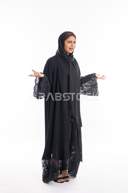 التعجب و الاستغراب من شيء ما، شابة سعودية تنظر الى الكاميرا بإستغراب، مفهوم النعومة والانوثة والاناقة، بورتريه لإمرأة عربية اماراتية خليجية مبتسمة محجبة ترتدي العباءة السوداء تقف باستقامة ترفع يديها نحو الأعلى بإيماءات الدهشة، الجسم بالطول الكامل، خلفية بيضاء