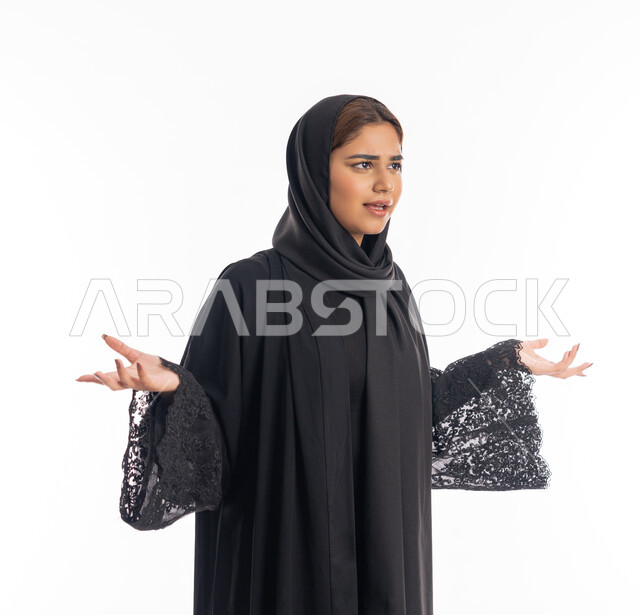 مفهوم الاناقة والنعومة والانوثة، الإندهاش و المفاجأة من شيء ما، شابة سعودية تنظر الى الكاميرا بإستغراب، بورتريه مقرب لإمرأة عربية اماراتية خليجية مبتسمة محجبة ترتدي العباءة السوداء تقف باستقامة ترفع يديها نحو الأعلى بإيماءات الدهشة، خلفية بيضاء