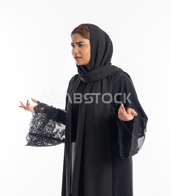 الإندهاش و المفاجأة من شيء ما، شابة سعودية تنظر الى الكاميرا بإستغراب، مفهوم النعومة والانوثة والاناقة، بورتريه مقرب لإمرأة عربية اماراتية خليجية مبتسمة محجبة ترتدي العباءة السوداء تقف باستقامة ترفع يديها نحو الأعلى بإيماءات الدهشة، خلفية بيضاء