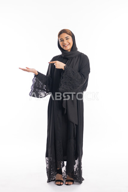 الاشارة إلى شيء ما والوقوف باستقامة، سيدة سعودية تنظر إلى الكاميرا بإيماءات السرور, تعابير التأكد وتحديد مكان معين, بورتريه لامرأة عربيه خليجيه إماراتية مبتسمة ترتدي العباءة السوداء ترفع اصبعها السبابة للاختيار، الجسم بالطول الكامل, خلفية بيضاء