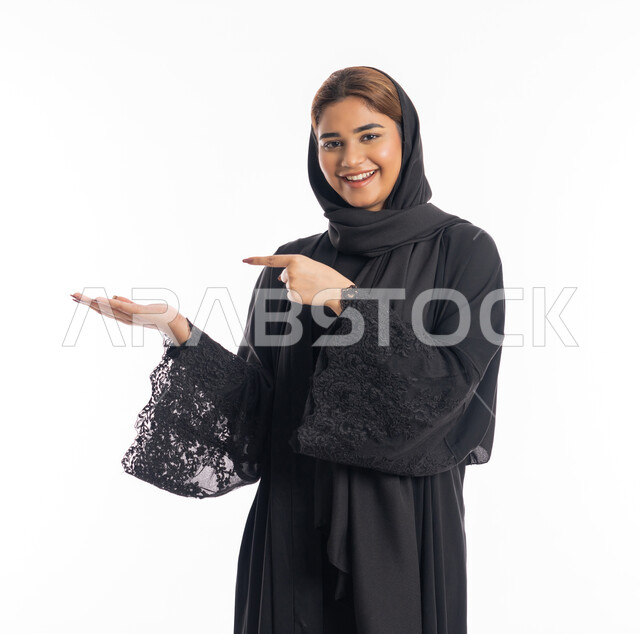 تعابير التأكد وتحديد مكان معين, الوقوف باستقامة والاشارة إلى شيء ما، سيدة سعودية تنظر إلى الكاميرا بإيماءات السرور, بورتريه مقرب لامرأة عربيه خليجيه إماراتية مبتسمة ترتدي العباءة السوداء ترفع اصبعها السبابة للاختيار, خلفية بيضاء