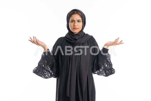 الاستغراب و التعجب من شيء ما، شابة سعودية تنظر الى الكاميرا بإستغراب، مفهوم النعومة والانوثة والاناقة، بورتريه مقرب لإمرأة عربية اماراتية خليجية مبتسمة محجبة ترتدي العباءة السوداء تقف باستقامة ترفع يديها نحو الأعلى بإيماءات الدهشة، خلفية بيضاء