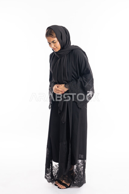 إيماءات التقيؤ والغثيان، الحاجه للمساعدة وطلب العلاج، الشعور بحرقة في المعدة وعسر الهضم، الإحساس بالمغص والوجع، سيدة عربية إماراتية ترتدي العباءة السوداء، بورتريه لامرأه خليجيه سعوديه تضع يدها على بطنها تنحني من شدة الألم، الجسم بالطول الكامل، خلفية بيضاء