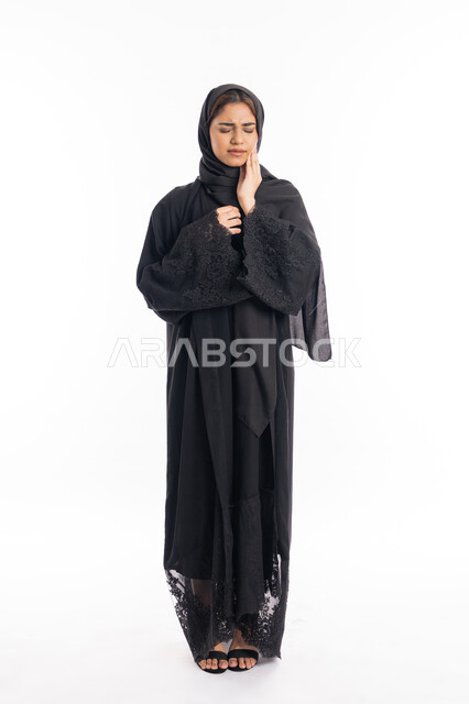 الحاجه لزيارة الطبيب، طلب العلاج و المساعده، سيدة عربية إماراتية ترتدي العباءة السوداء، تسوس الأضراس وإلتهاب اللثه، بورتريه لامرأه خليجيه سعوديه محجبة تضع يدها على خدها بإيماءات الشعور بألم الأسنان، الجسم بالطول الكامل، خلفية بيضاء