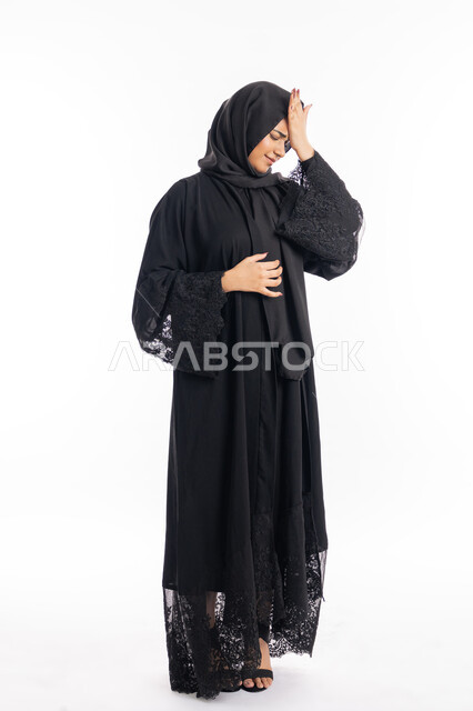 Expressions of fatigue and exhaustion, feeling head pain and headache, close-up portrait of a veiled Saudi Gulf woman placing her hands on her head with gestures of suffering from migraine pain, the need for calm and rest, seeking treatment and help, an Emirati Arab woman wearing a black abaya, white background.