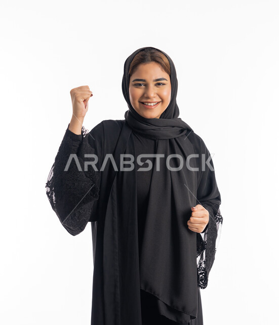 The concept of joy in victory and victory, achieving goals and success, an Emirati Arab woman wearing a black abaya looking at the camera with expressions of pleasure and happiness, a close-up portrait of a veiled Saudi Gulf woman, smiling, raising her hands with enthusiastic gestures, white background.
