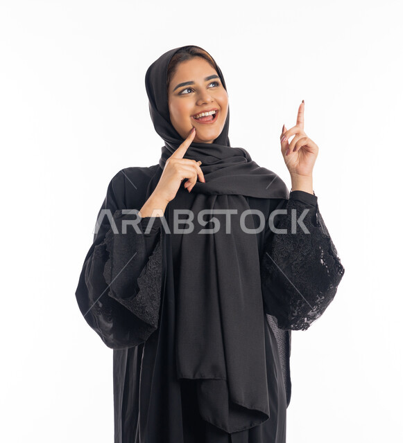 الوقوف والنظر إلى شيء ما, بورتريه مقرب  لامرأه خليجيه سعوديه مبتسمة ترفع اصبع السبابة لجهة اليسار لتعين موقع محدد, الإشارة لمكان ما بإيماءات التخطيط, التأمل والتفكير بالأهداف المستقبلية, سيدة عربية إماراتية ترتدي العباءة السوداء، خلفية بيضاء