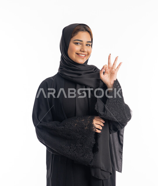 تعابيرالرضى و الاعجاب، علامة الامتياز و الجودة، بورتريه مقرب لامراة عربية خليجية اماراتية مبتسمة محجبة ترتدي العباءة السوداء  ترفع يدها بإيماءات تدل على القبول و الموافقة، خلفية بيضاء