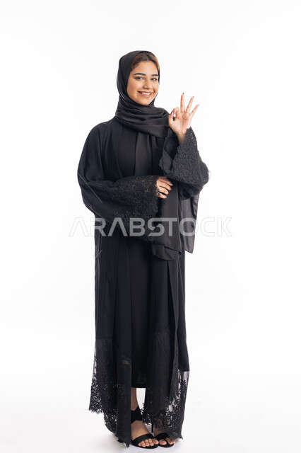 مفهوم القبول والموافقه، علامة الامتياز و الجودة، بورتريه لامراة عربية خليجية اماراتية مبتسمة محجبة ترتدي العباءة السوداء  ترفع يدها بإيماءات تدل على الإعجاب والرضى، الجسم بالطول الكامل، خلفية بيضاء