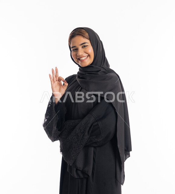 تعابير الإعجاب والرضى، علامة الامتياز و الجودة، بورتريه مقرب لامراة عربية خليجية اماراتية مبتسمة محجبة ترتدي العباءة السوداء  ترفع يدها بإيماءات تدل على القبول و الموافقة، خلفية بيضاء