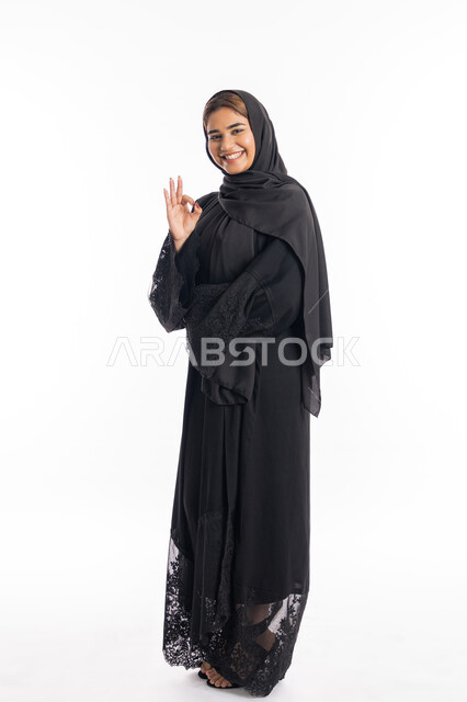 علامة الامتياز و الجودة، مفهوم القبول والموافقه، بورتريه لامراة عربية خليجية اماراتية مبتسمة محجبة ترتدي العباءة السوداء  ترفع يدها بإيماءات تدل على الإعجاب والرضى، الجسم بالطول الكامل، خلفية بيضاء